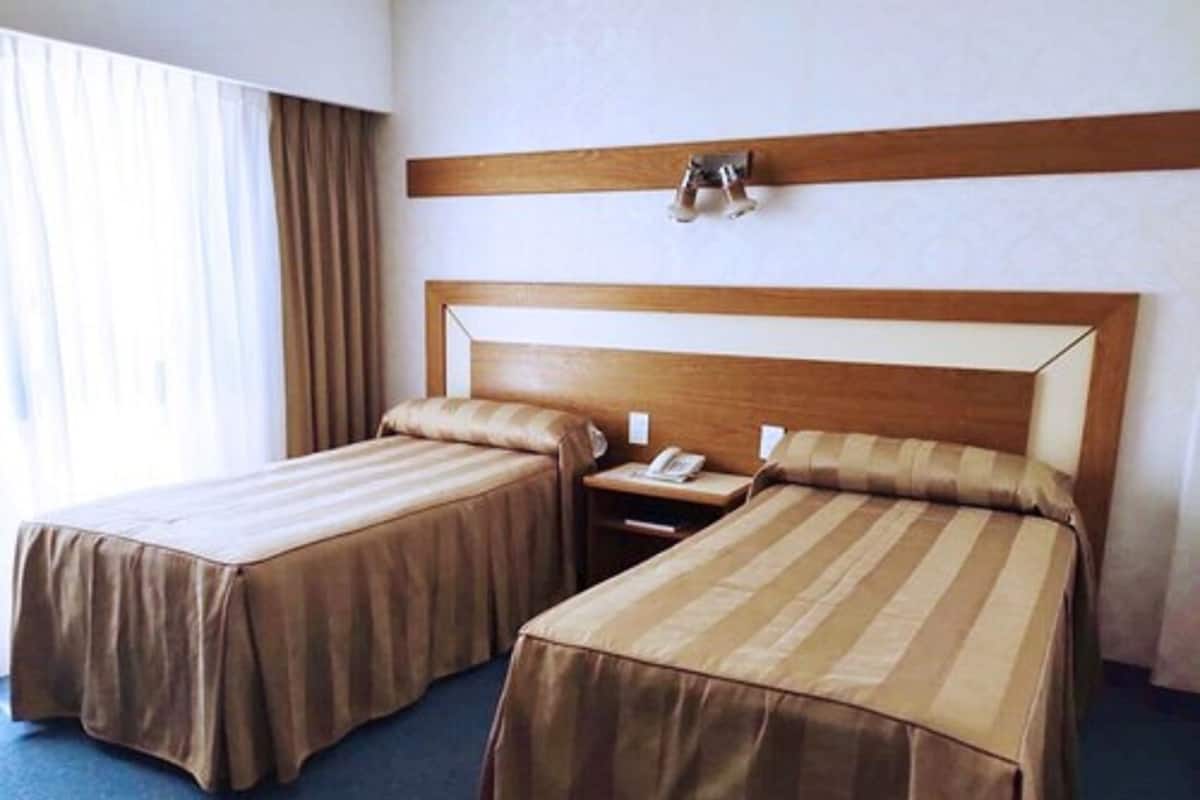 A hotel room with two single beds, a wooden headboard, a bedside table with a phone, and a window with curtains.