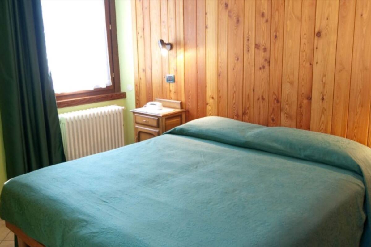 Ein Zimmer mit einem Bett, einem kleinen Nachttisch, einem Heizkörper und einem Fenster mit Vorhängen.