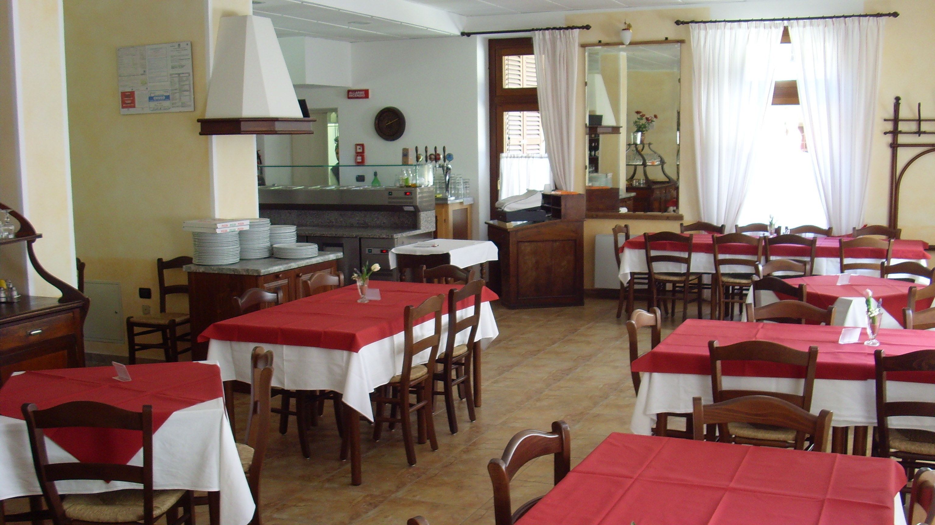 Ein Restaurant mit mehreren Tischen, die mit roten Tischdecken bedeckt sind, Holzsesseln und einer Küche im Hintergrund.