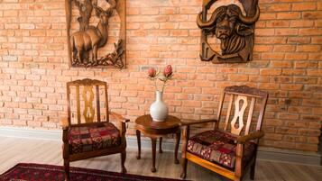 Una habitación con pared de ladrillo, dos sillas de madera con cojines estampados, una pequeña mesa redonda de madera con un jarrón blanco, y un tapete rojo con diseño en el suelo.