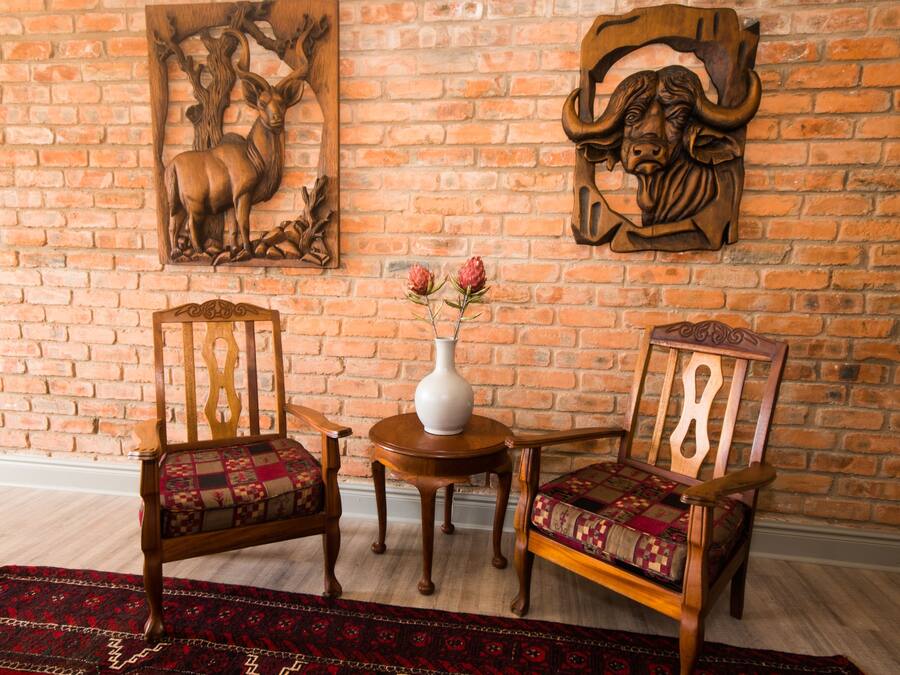 Una habitación con pared de ladrillo, dos sillas de madera con cojines estampados, una pequeña mesa redonda de madera con un jarrón blanco, y un tapete rojo con diseño en el suelo.