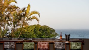 Eine Bar mit gemusterten Stühlen und einer Flasche, mit Blick auf ein Gewässer und Palmen.