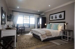 Un dormitorio con cama, mesitas de noche, escritorio, silla, un ventanal con cortinas y una obra de arte en la pared.