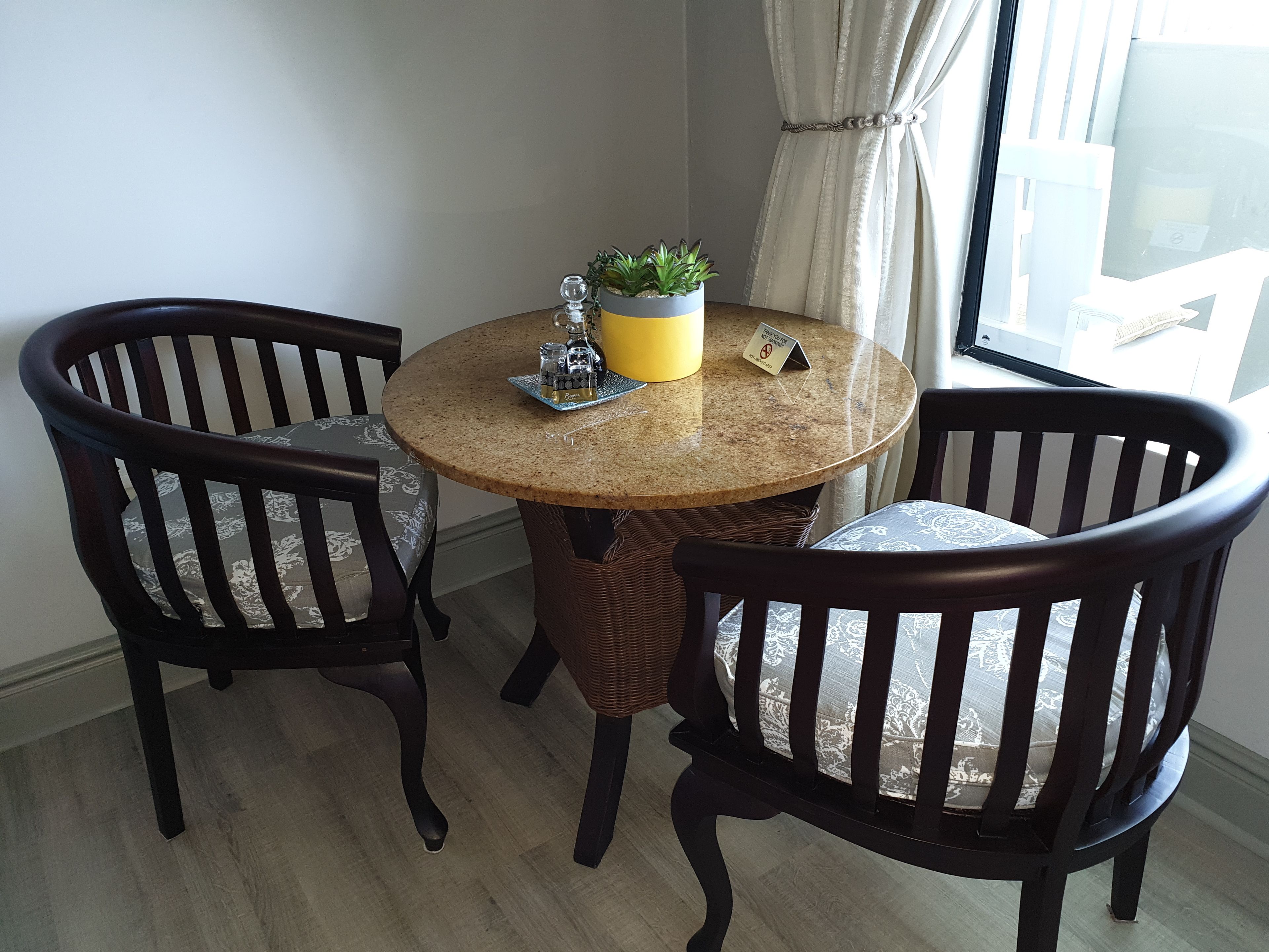 A round table with a woven base surrounded by four dark wooden chairs with patterned cushions. A small potted plant and a yellow mug are on the table.