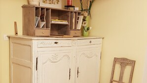 Una habitación con un armario blanco y ornamentado, con múltiples puertas y cajones, una silla de mimbre y un jarrón con flores encima del armario.