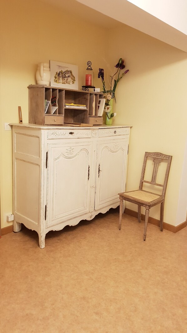 A room with a white, ornate cabinet with multiple doors and drawers, a wicker chair, and a vase with flowers on top of the cabinet. - Au Bel Air (Avallon)