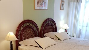 Una cama bien tendida con una colcha de estampado floral, dos mesitas de noche con lámparas, un cuadro en la pared y una ventana con cortinas.