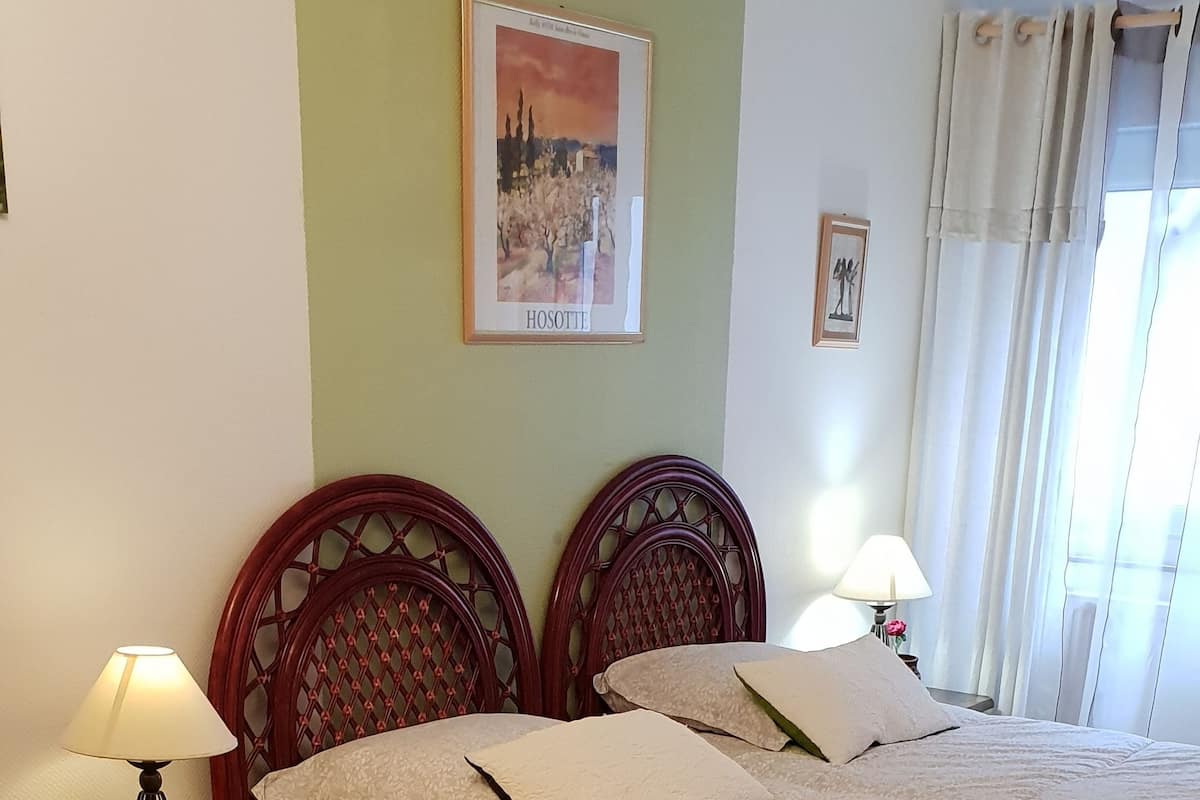 Una cama bien tendida con una colcha de estampado floral, dos mesitas de noche con lámparas, un cuadro en la pared y una ventana con cortinas.