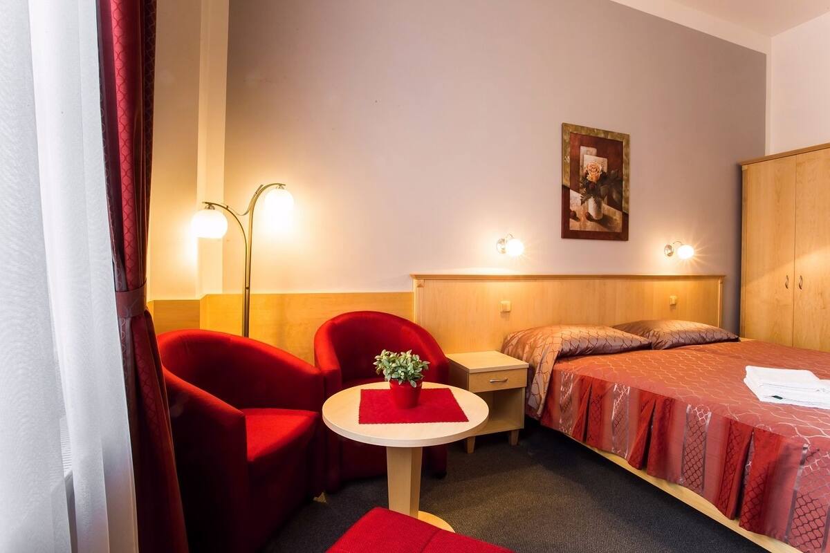 Habitación de hotel con una cama, dos sillas rojas, una mesita con una planta y un cuadro en la pared.