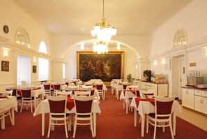 Une salle à manger élégante, avec des tables blanches et des chaises rouges, un grand tableau au mur et un lustre.