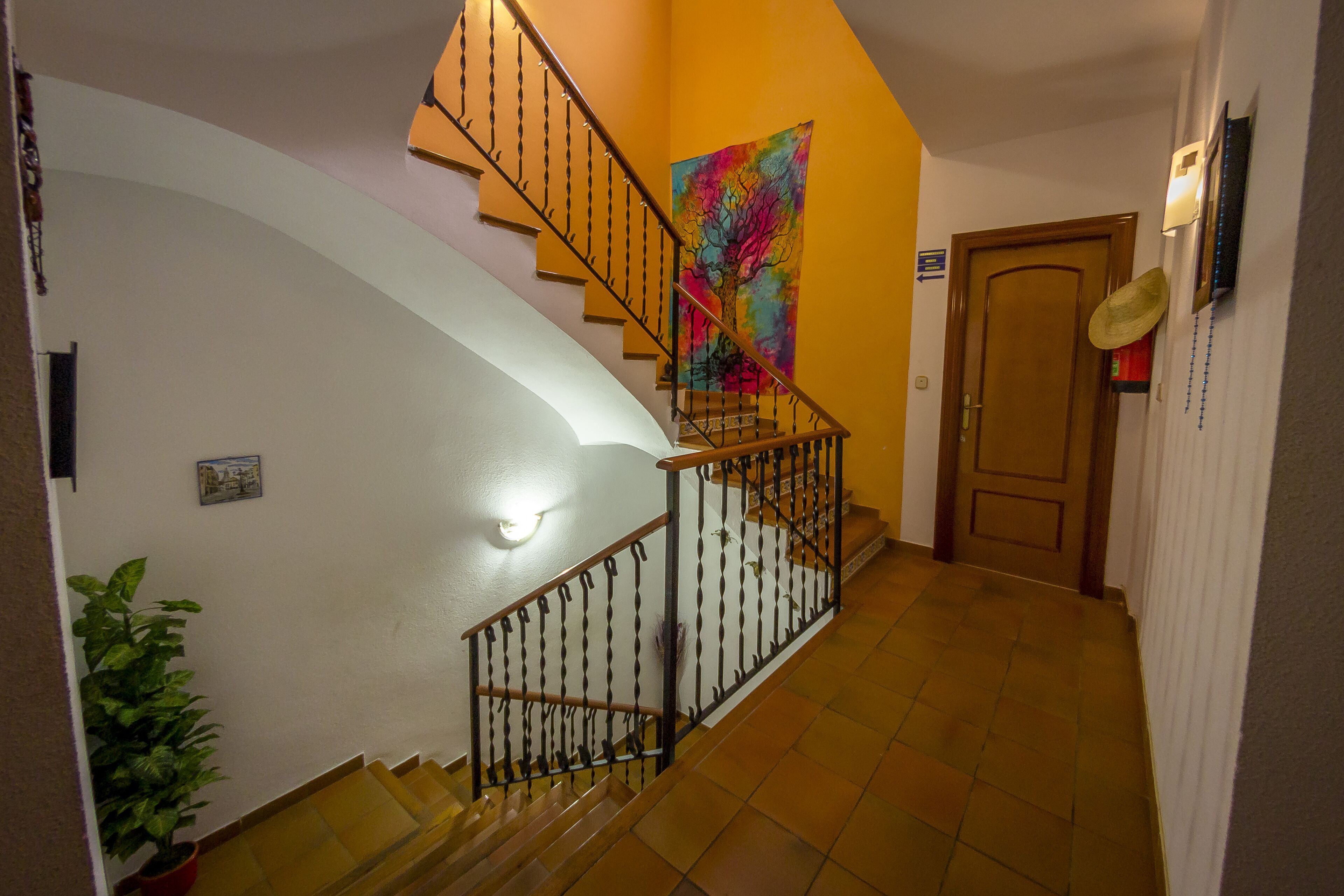 Una escalera con barandilla metálica, un tapiz de colores en la pared, una planta en maceta y una puerta con lámpara.
