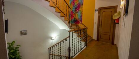 Una escalera con barandilla metálica, un tapiz de colores en la pared, una planta en maceta y una puerta con lámpara.