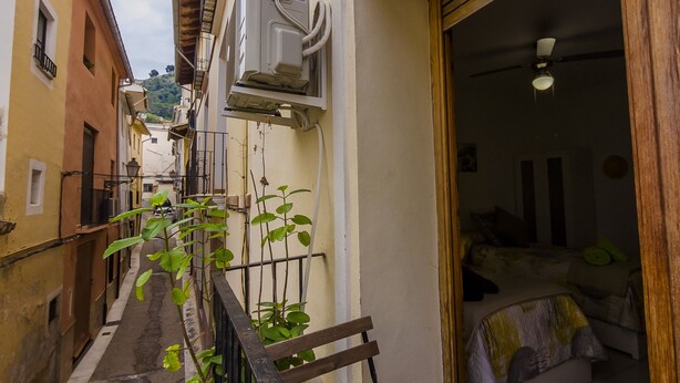 Un estrecho callejón con un banco de madera y una barandilla de metal. A la derecha, un edificio con un balcón y una puerta que da a una habitación con cama y ventilador de techo.