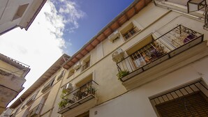Una calle estrecha con edificios que tienen balcones con plantas, una puerta visible y un cielo azul claro.