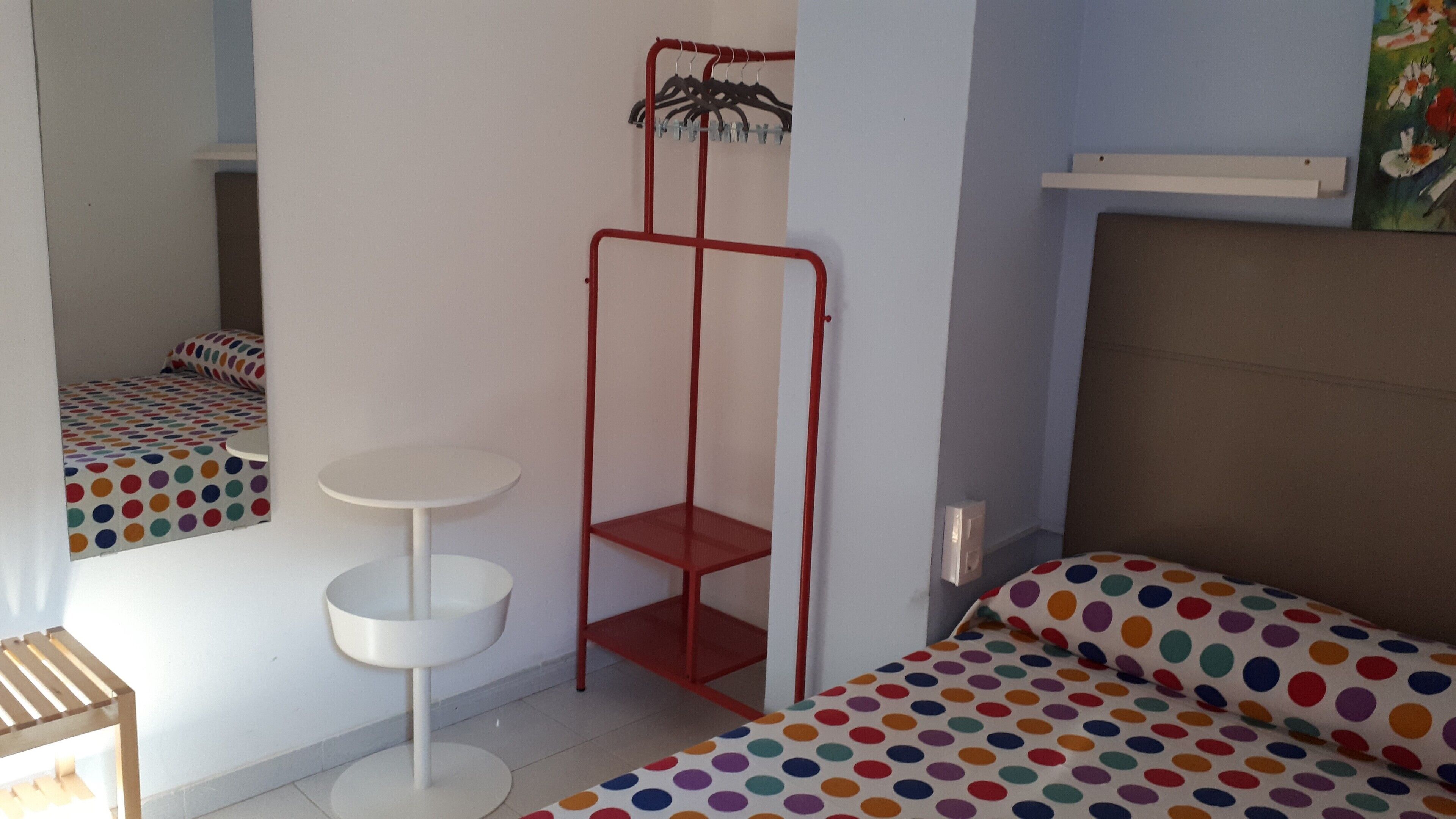 A room with a bed, a small white table, a red metal shelf, and a colorful polka dot bedspread.