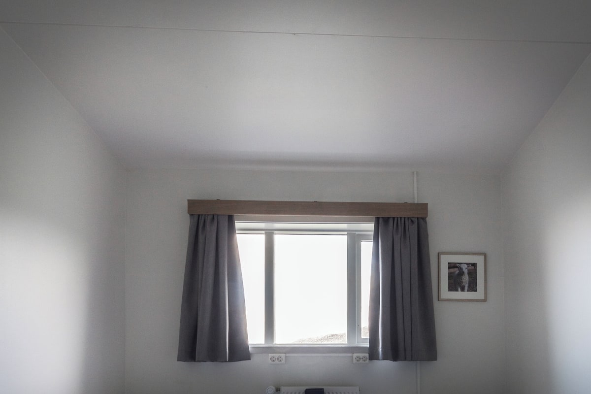 A small, well-lit room with two single beds, a radiator, a small table, and a window with curtains.
