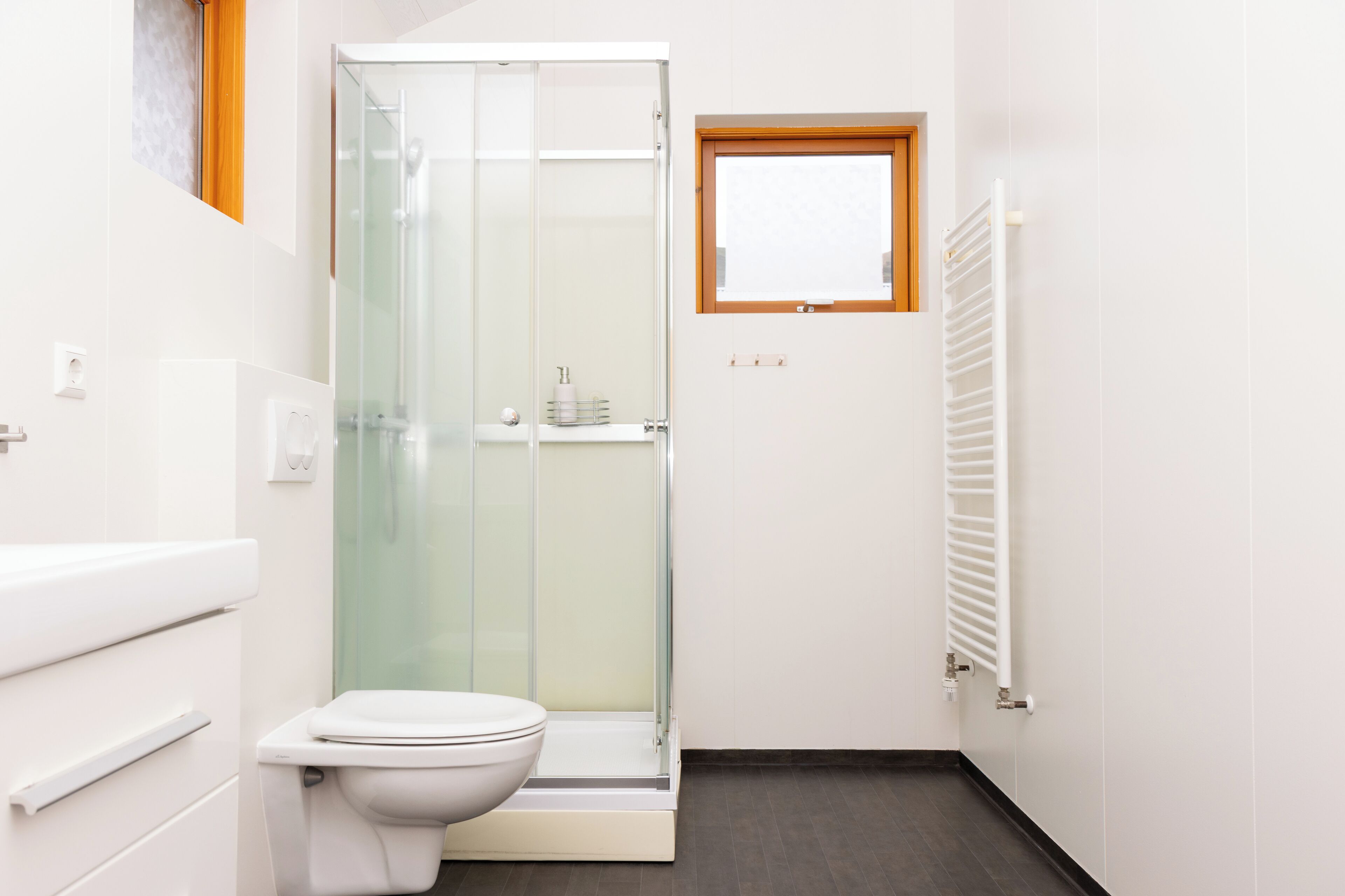A modern bathroom with a glass shower enclosure, a toilet, a small window, and a white radiator.