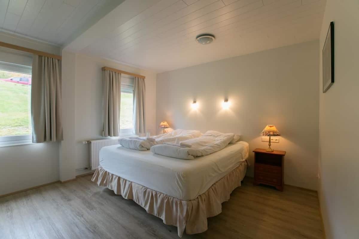 A bedroom with a bed, two bedside lamps, a window with curtains, and a radiator.
