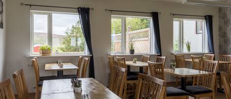 A dining area with multiple wooden tables and chairs, large windows with curtains, and a view of the outdoors.