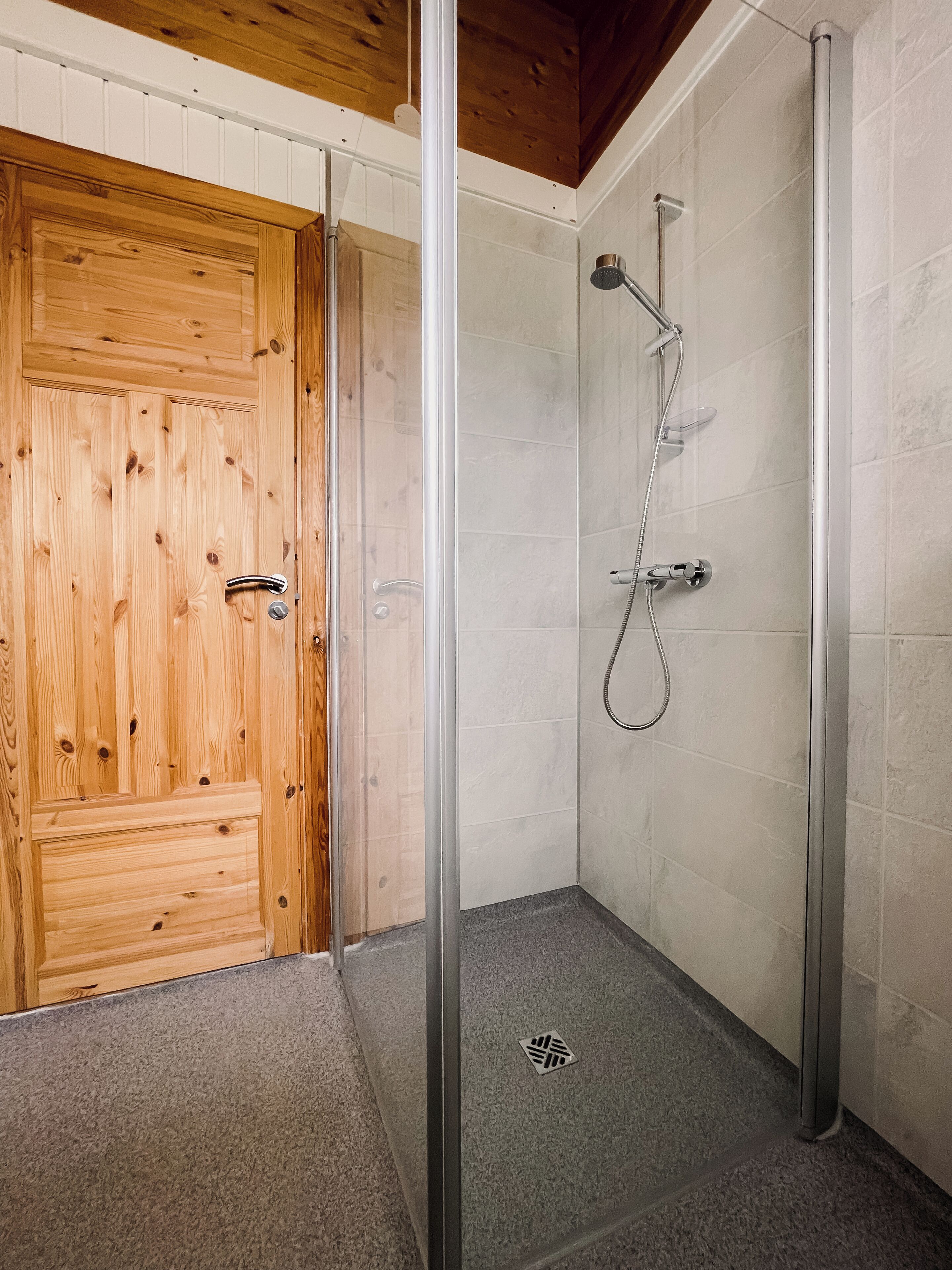 A wooden door with a metal handle, a glass shower enclosure with a showerhead and handheld shower, and a small drain in the floor.