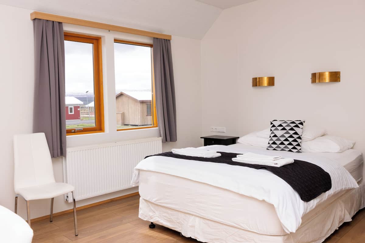 A bedroom with a bed, a chair, a window with curtains, a radiator, and a view of a building outside.
