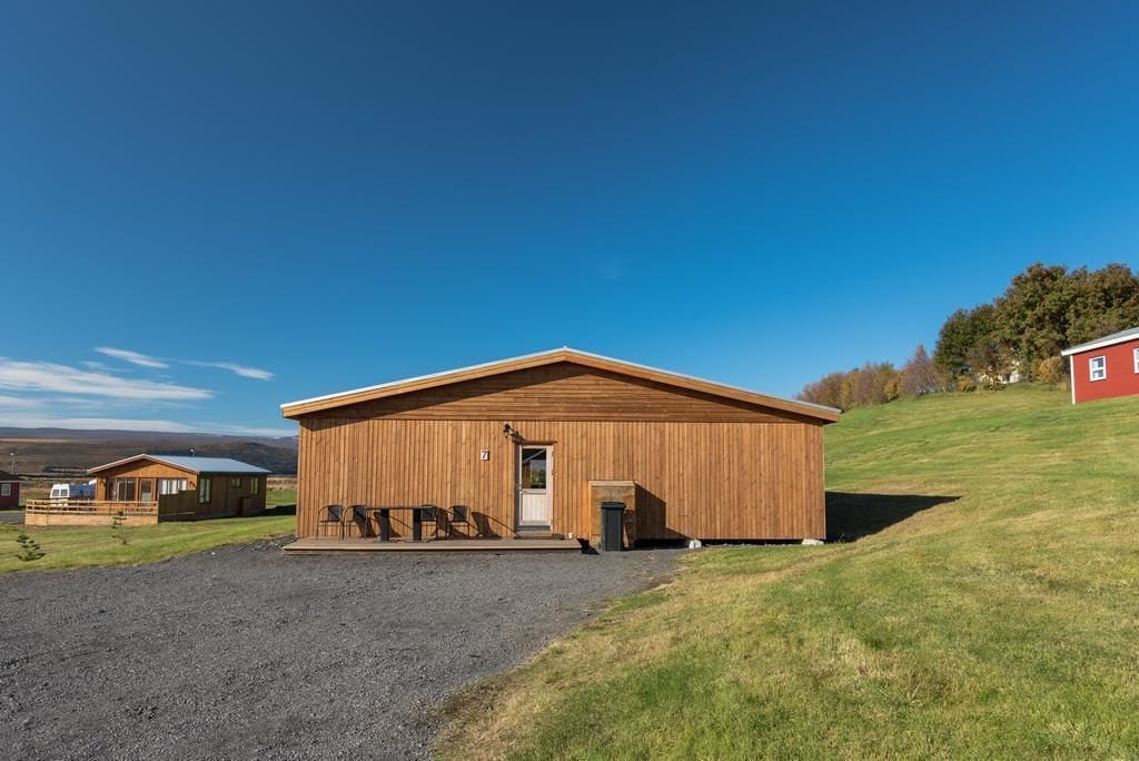 A wooden cabin with a porch and outdoor seating, surrounded by a gravel driveway and grassy area.