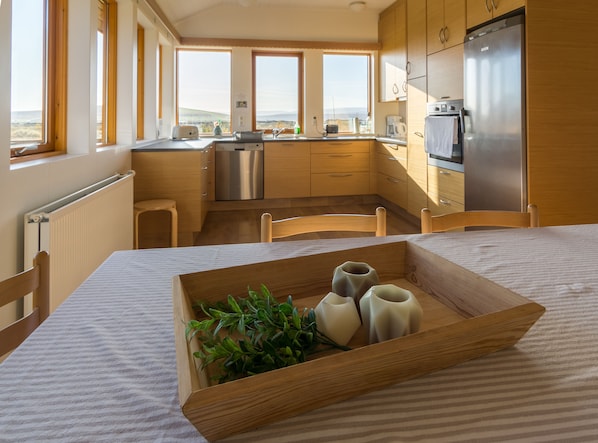 A modern kitchen with wooden cabinets, a stainless steel refrigerator, and a view of the landscape through large windows. - Guesthouse Brekka (Husavik)