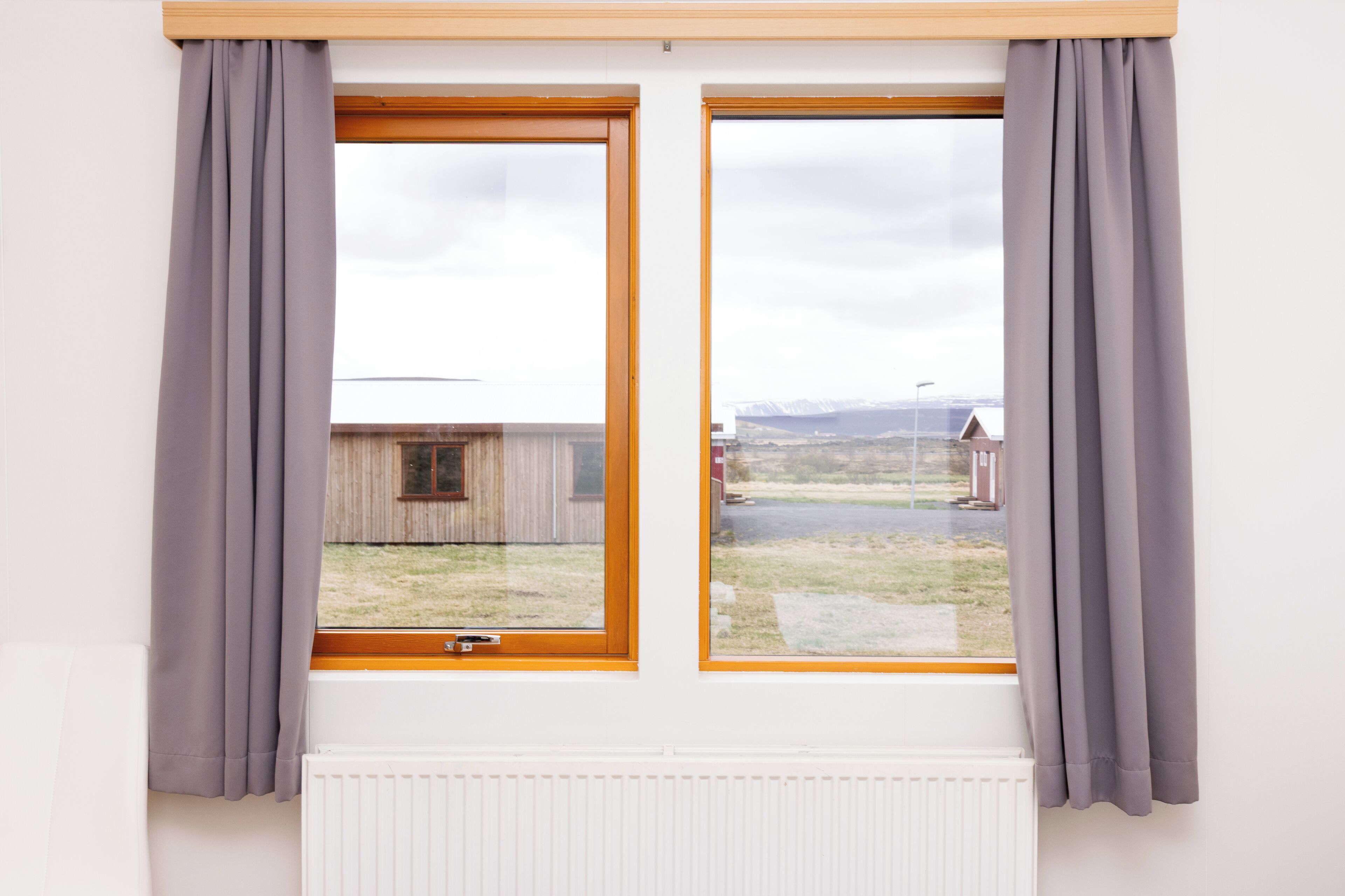 A window with wooden frames and grey curtains, overlooking a grassy area with a wooden building and a distant mountain range.