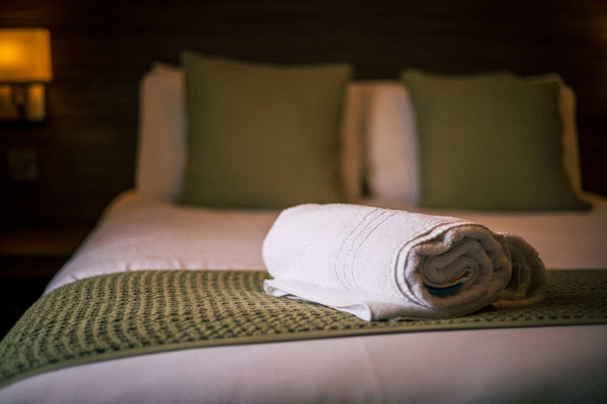 Una cama bien hecha con una toalla blanca enrollada, un plaid verde y dos almohadas verdes.