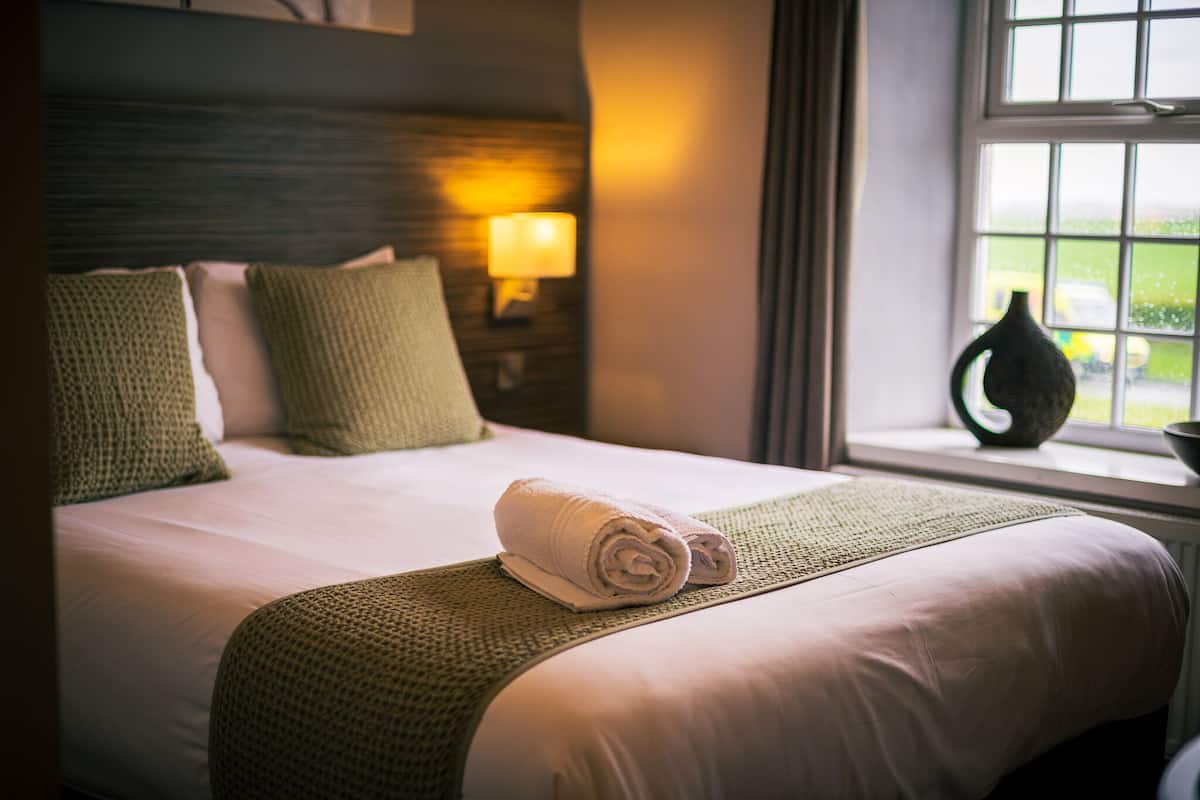 Uma cama bem arrumada com uma toalha enrolada, um cobertor verde e uma luminária na mesa de cabeceira.