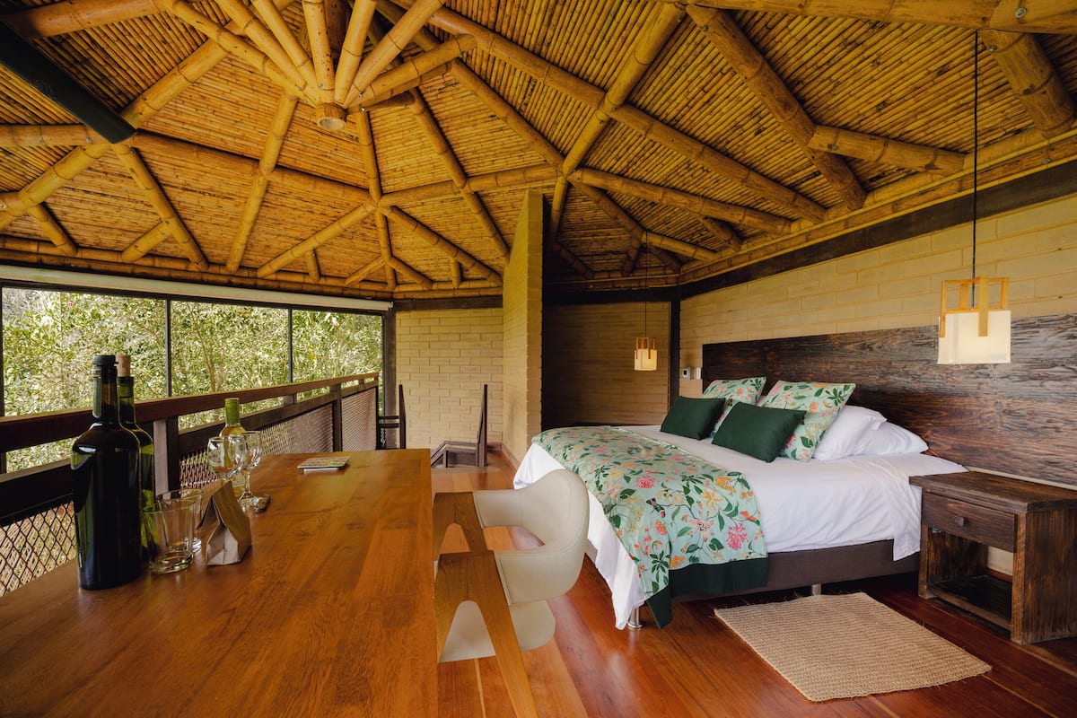 A room with a wooden ceiling, a bed with floral bedding, a wooden table with glasses and a bottle, and a view through large windows.