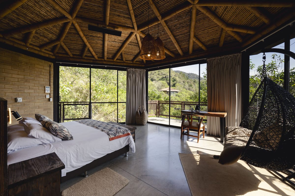 A bedroom with a large bed, a desk, and a hanging chair, with a view of greenery outside.