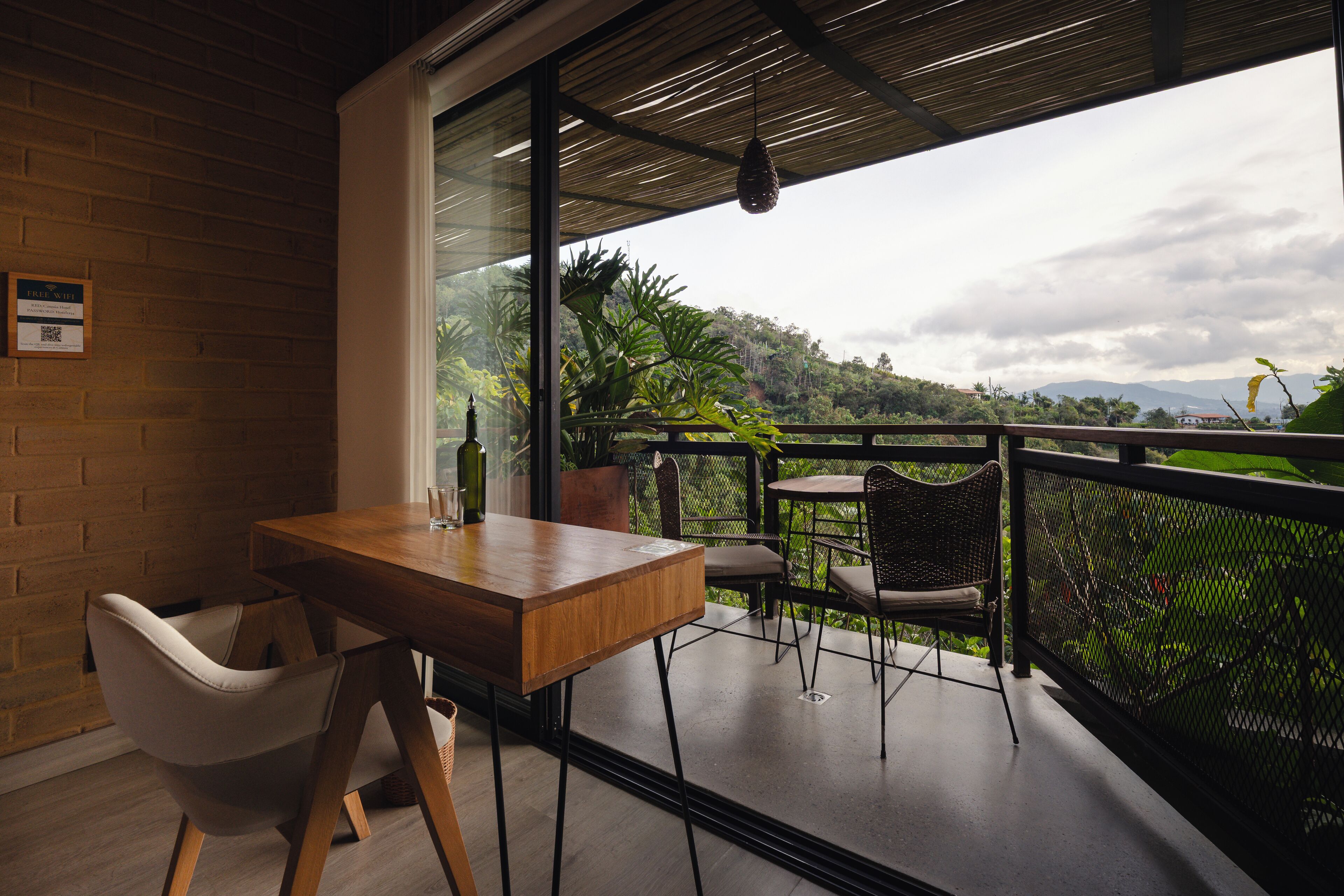 A balcony with a wooden table and chairs, a glass door leading to a lush garden, and a view of distant mountains.
