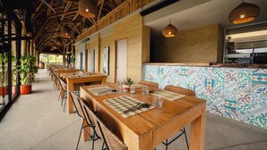 Un comedor amplio con mesas y sillas de madera, una zona de cocina con salpicadero de azulejos y grandes ventanales que dejan entrar luz natural.