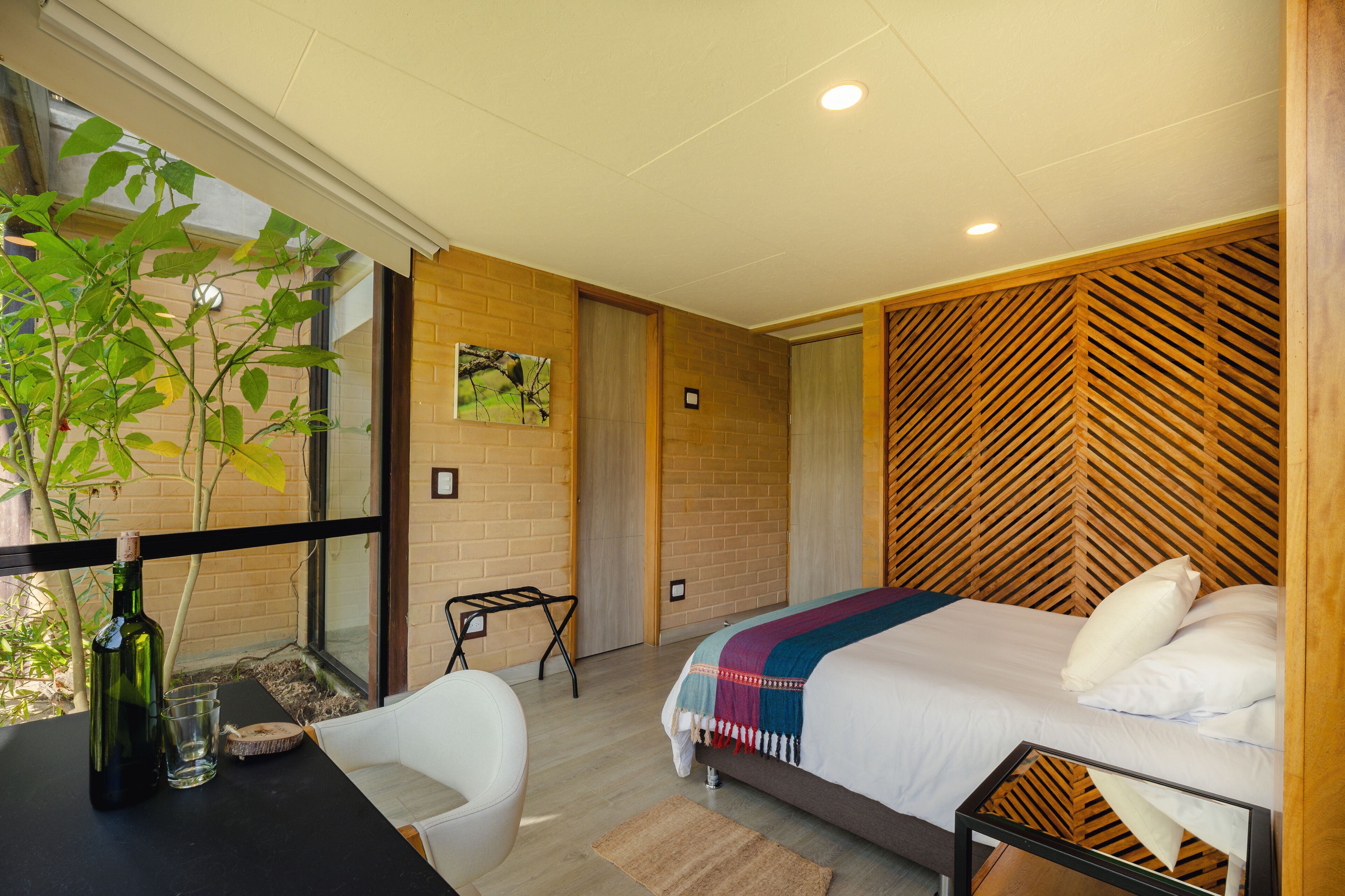 A bedroom with a bed, a desk with a bottle and glass, a chair, a window with a plant, and a wooden wall panel.