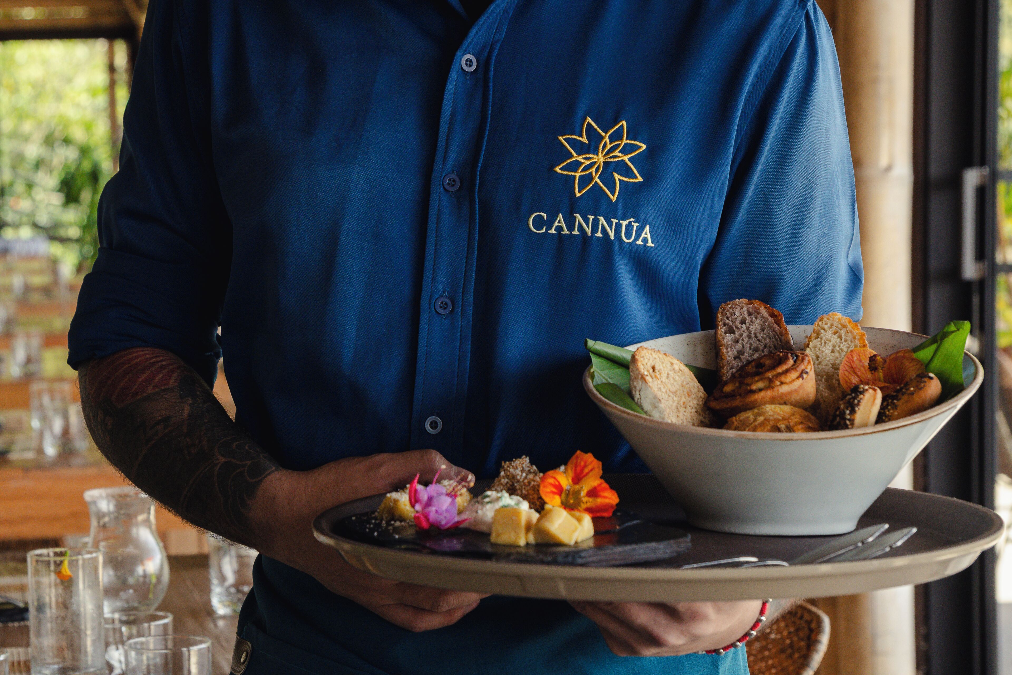 Una persona con una camicia blu con un logo che tiene un vassoio con una ciotola di pane e un piccolo piatto di cibo.