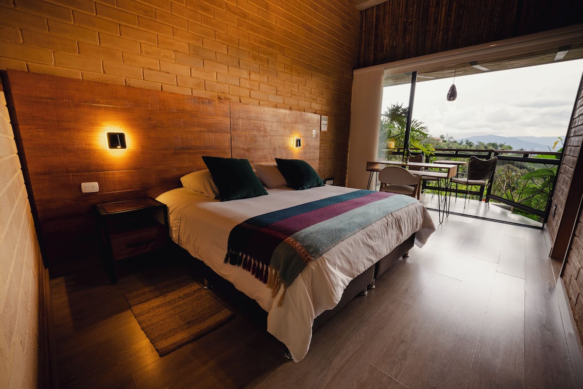 A bedroom with a large bed, a nightstand, a table with chairs, and a balcony with a view.
