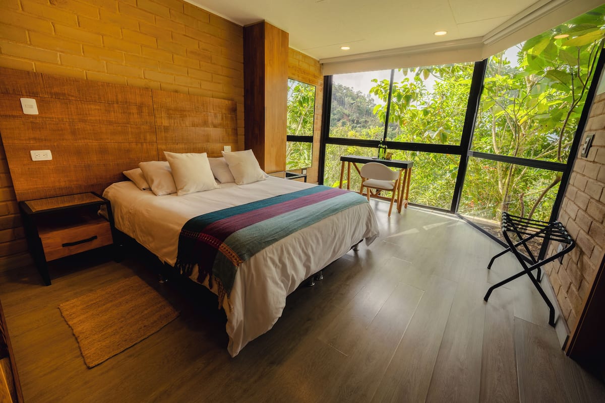A bedroom with a large bed, a wooden nightstand, a small table with a chair, and a view of greenery through large windows.