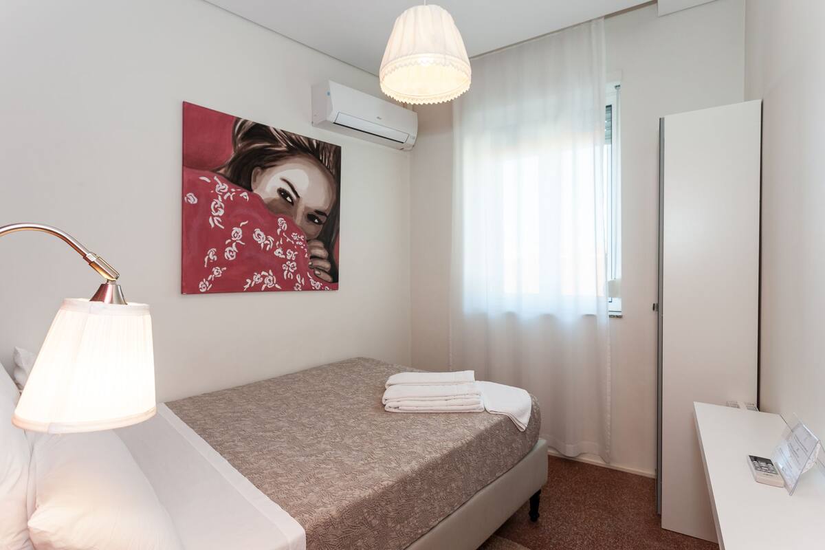 Ein Hotelzimmer mit Bett, Nachttisch, Lampe, einem Bild an der Wand und einer Klimaanlage.