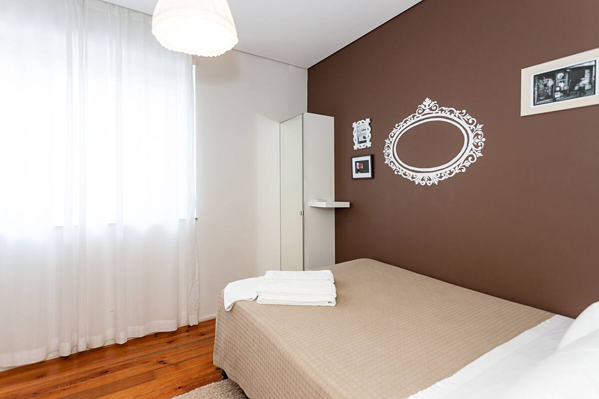 Ein Schlafzimmer mit einem Bett, einem großen Spiegel und einem Fenster mit durchsichtigen Vorhängen.