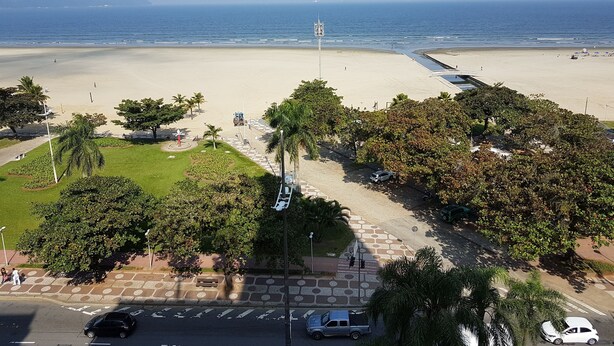 A beach with a clear view of the ocean, a parking area, and a pedestrian path. - Apartamento em frente a Praia Santos (Santos)