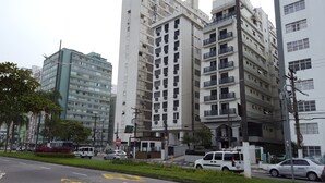 A multi-story residential building with balconies, a small entrance with a sign, and a street with cars and a bus. - Apartamento em frente a Praia Santos (Santos)