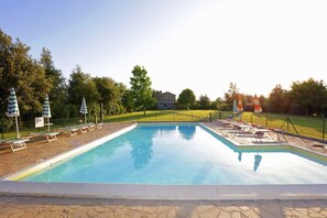 A rectangular swimming pool with lounge chairs and umbrellas, surrounded by a stone deck and a grassy area with trees. - Agriturismo Buriano (Lubriano)