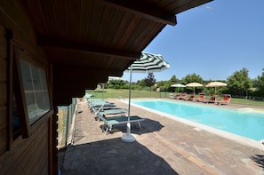 A pool area with lounge chairs and umbrellas, a stone patio, and a wooden building with a window.