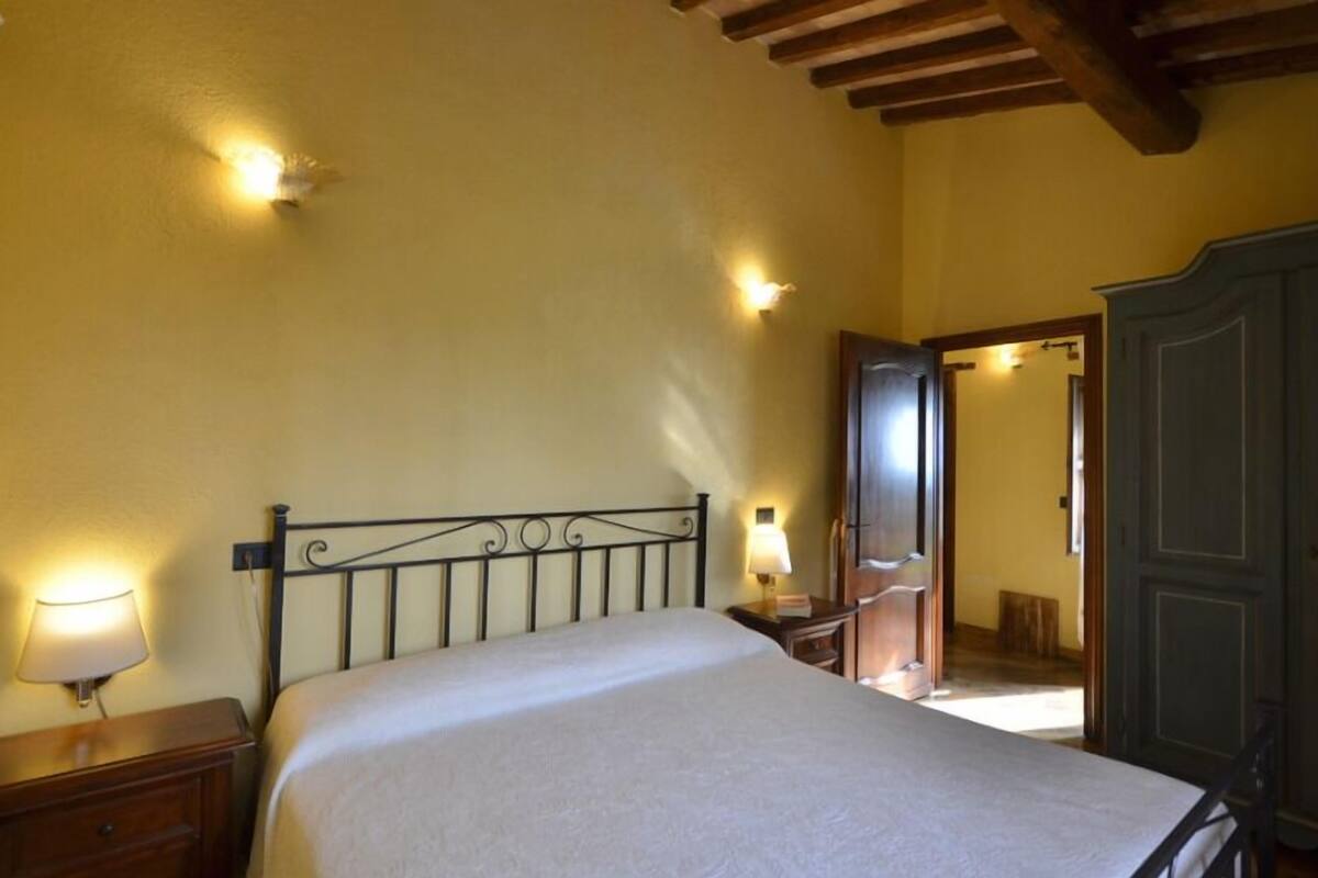 A bedroom with a bed, bedside table, lamp, wardrobe, and a door leading to another room.