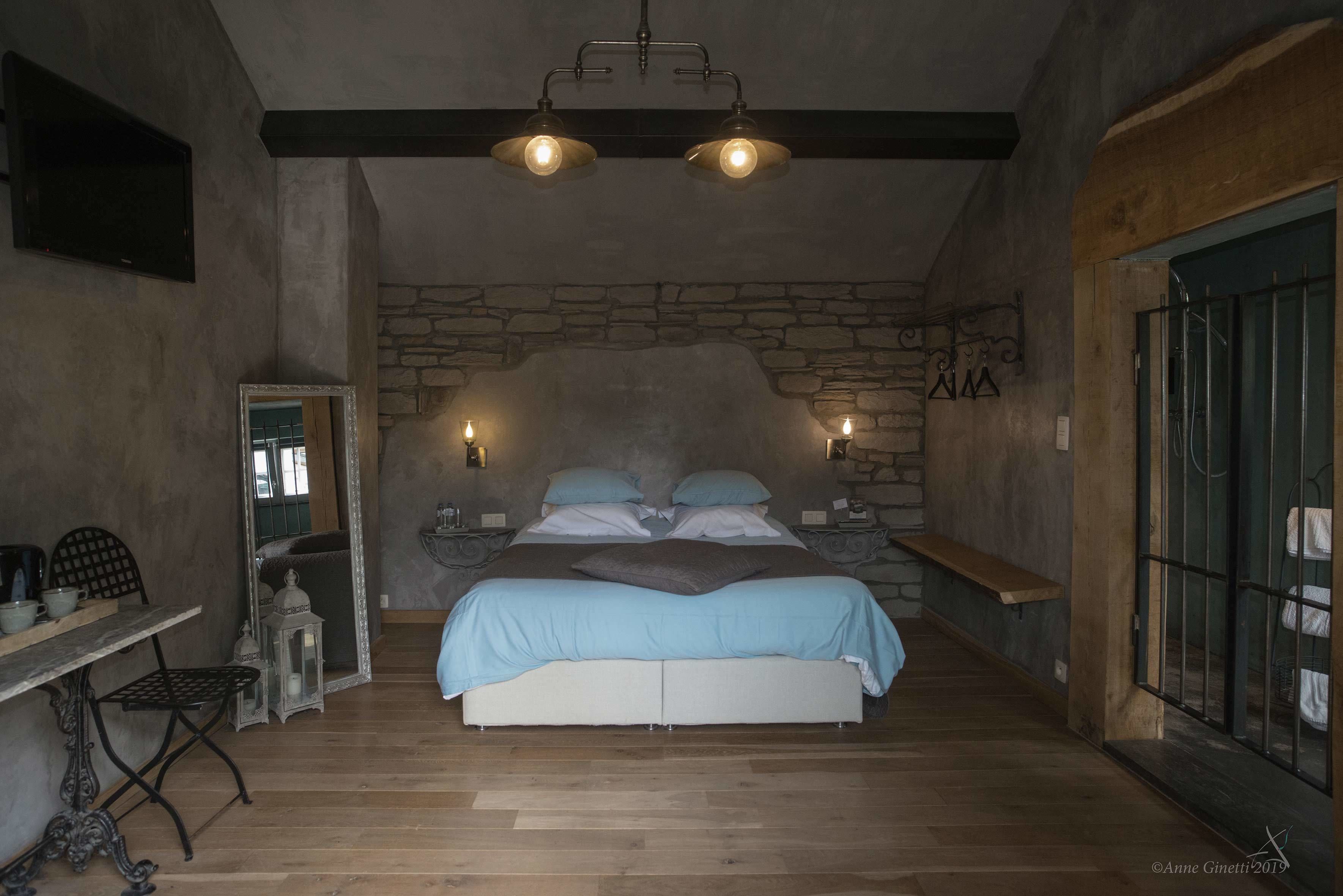 Un dormitorio con pared de piedra, cama con colcha azul claro, dos lámparas de noche, una mesita de noche, un banco de madera y una pequeña mesa con una silla.