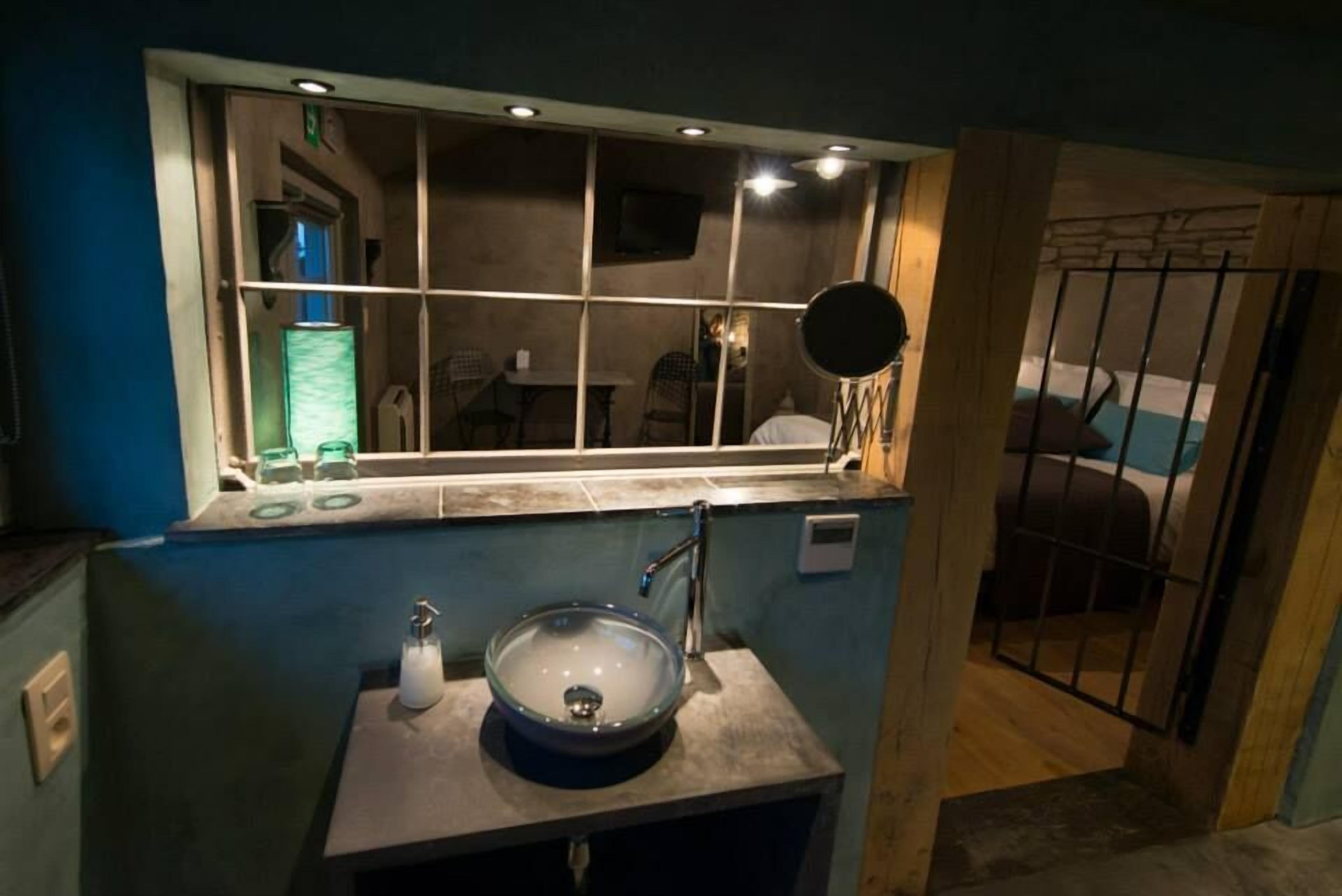 A bathroom with a glass sink, a mirror, and a window with a view of a room.