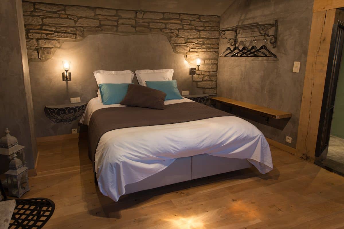 Un dormitorio con pared de piedra, una cama con ropa de cama blanca y azul, dos mesitas de noche con lámparas y un banco de madera.