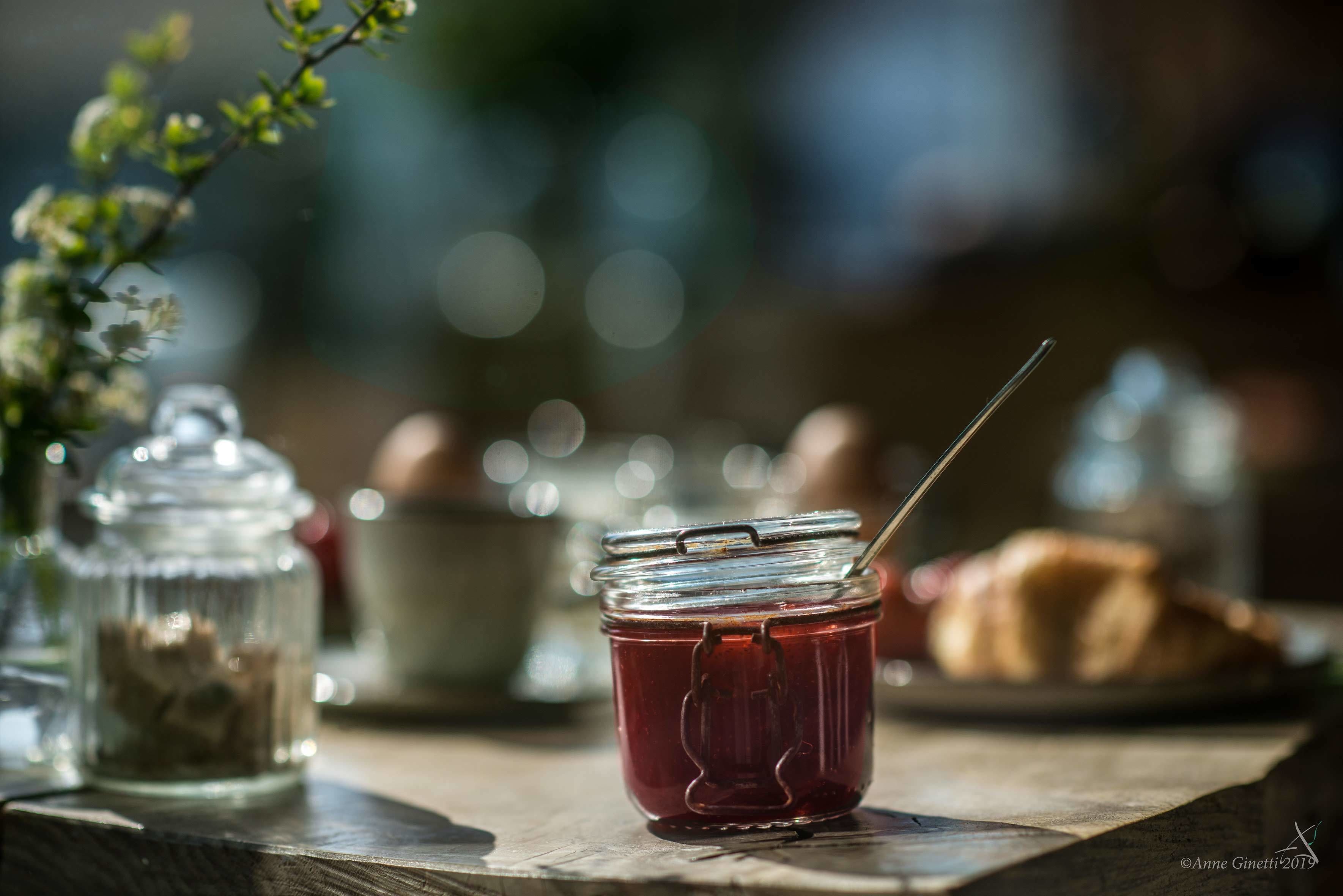 Een pot jam met een lepel op een houten tafel, een glazen pot met deksel, en een gebakje op de achtergrond.