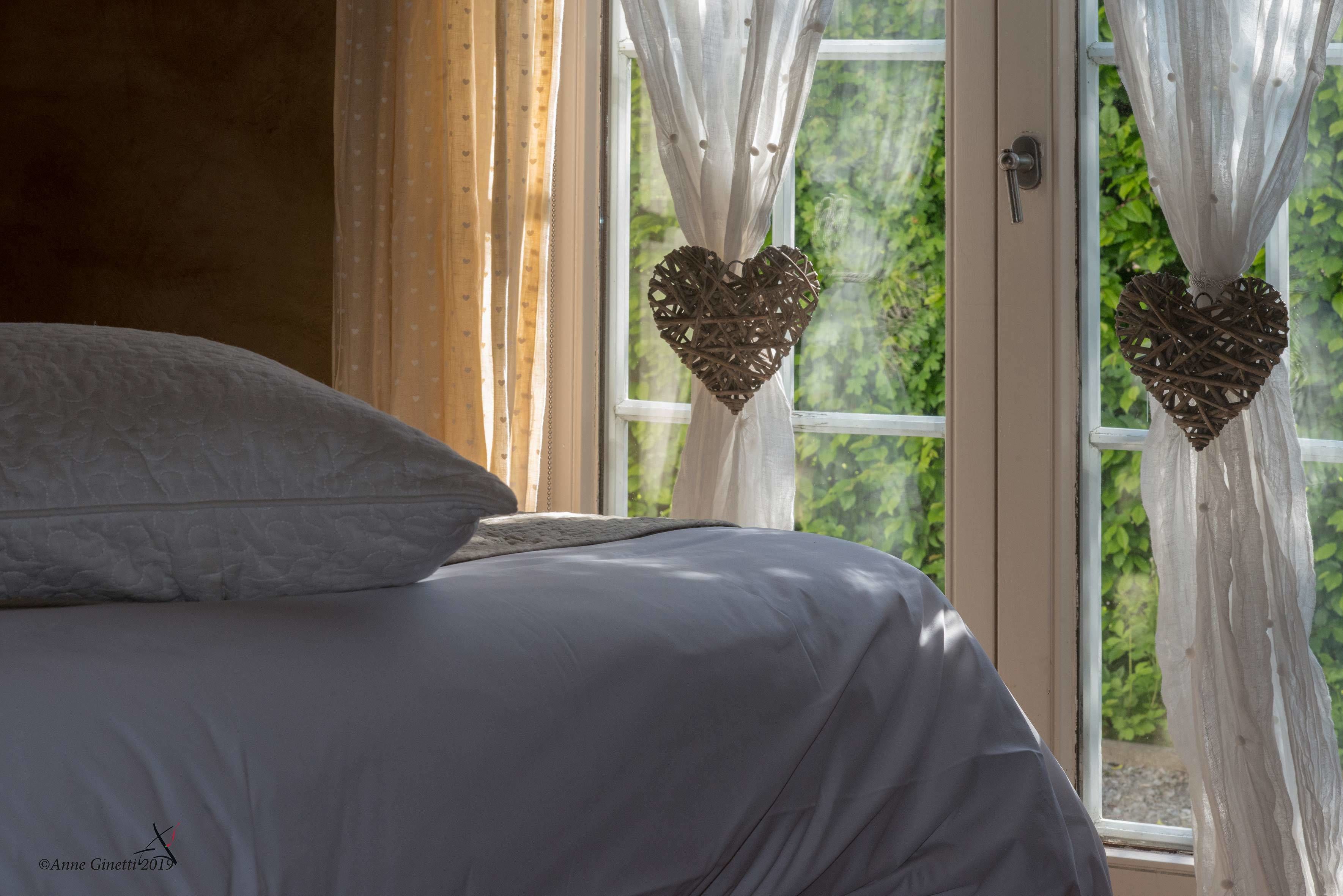Ein Bett mit weißer Bettwäsche, ein Fenster mit Vorhängen und zwei herzförmige Dekorationen.